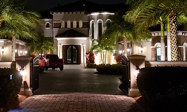 Well-lit paver driveway approach to an expensive home surrounded by up-lit palm trees and vegetation.