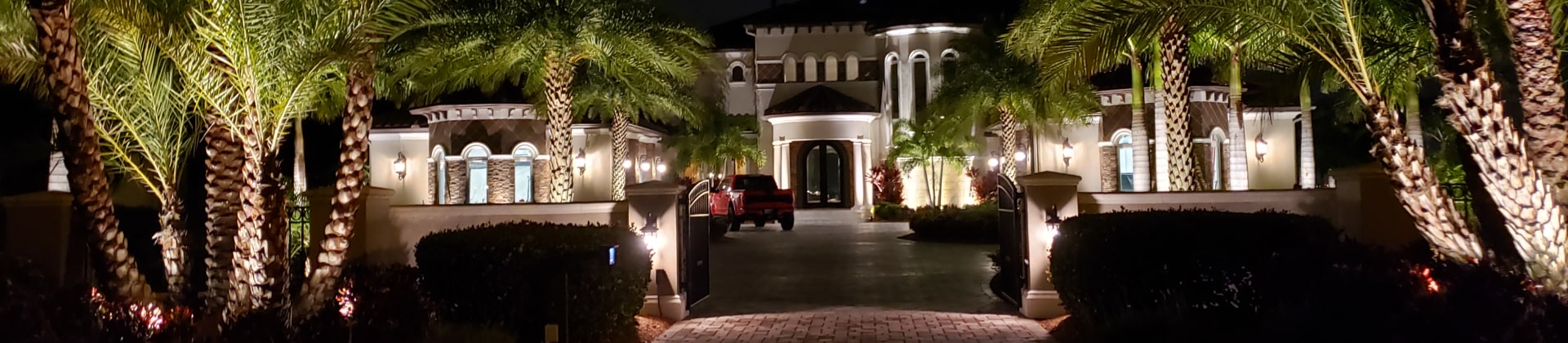 Well-lit Paver Driveway Approach To An Expensive Home Surrounded By Up-lit Palm Trees and Vegetation.